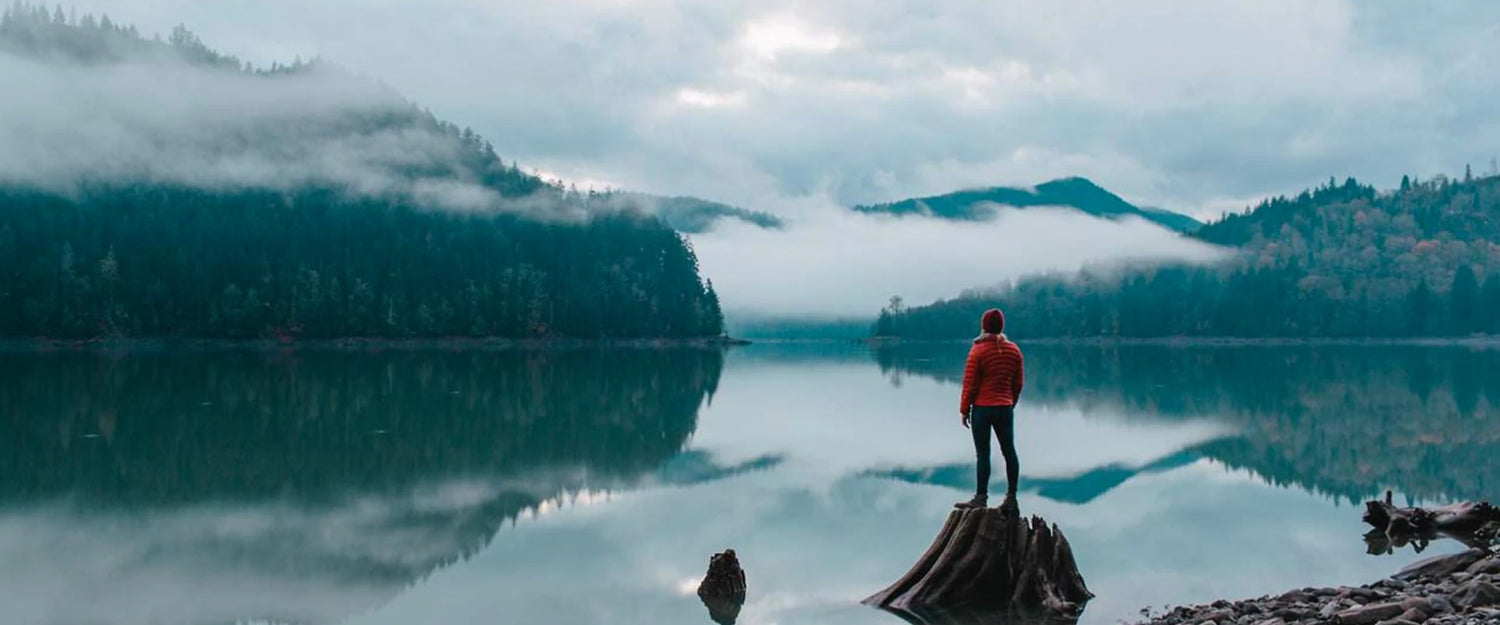 A person in a red jacket stands on a log by a calm lake surrounded by misty mountains and forested hills under a cloudy sky.