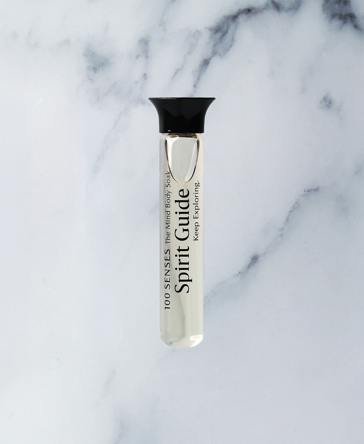 A small cylindrical glass vial with a black cap, labeled 'Spirit Guide: Keep Exploring' and '100 SENSES The Mind Body Soak', placed on a white marble surface.