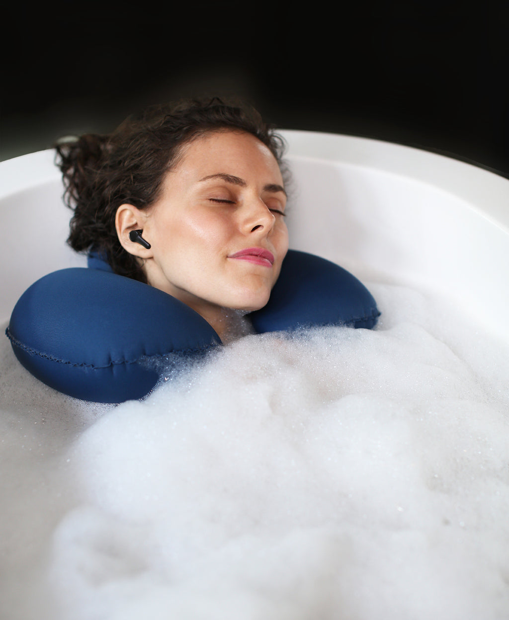 Person wearing BathSonic earbuds while resting in a foaming bath soak with a blue Head Floater snapped around their neck.