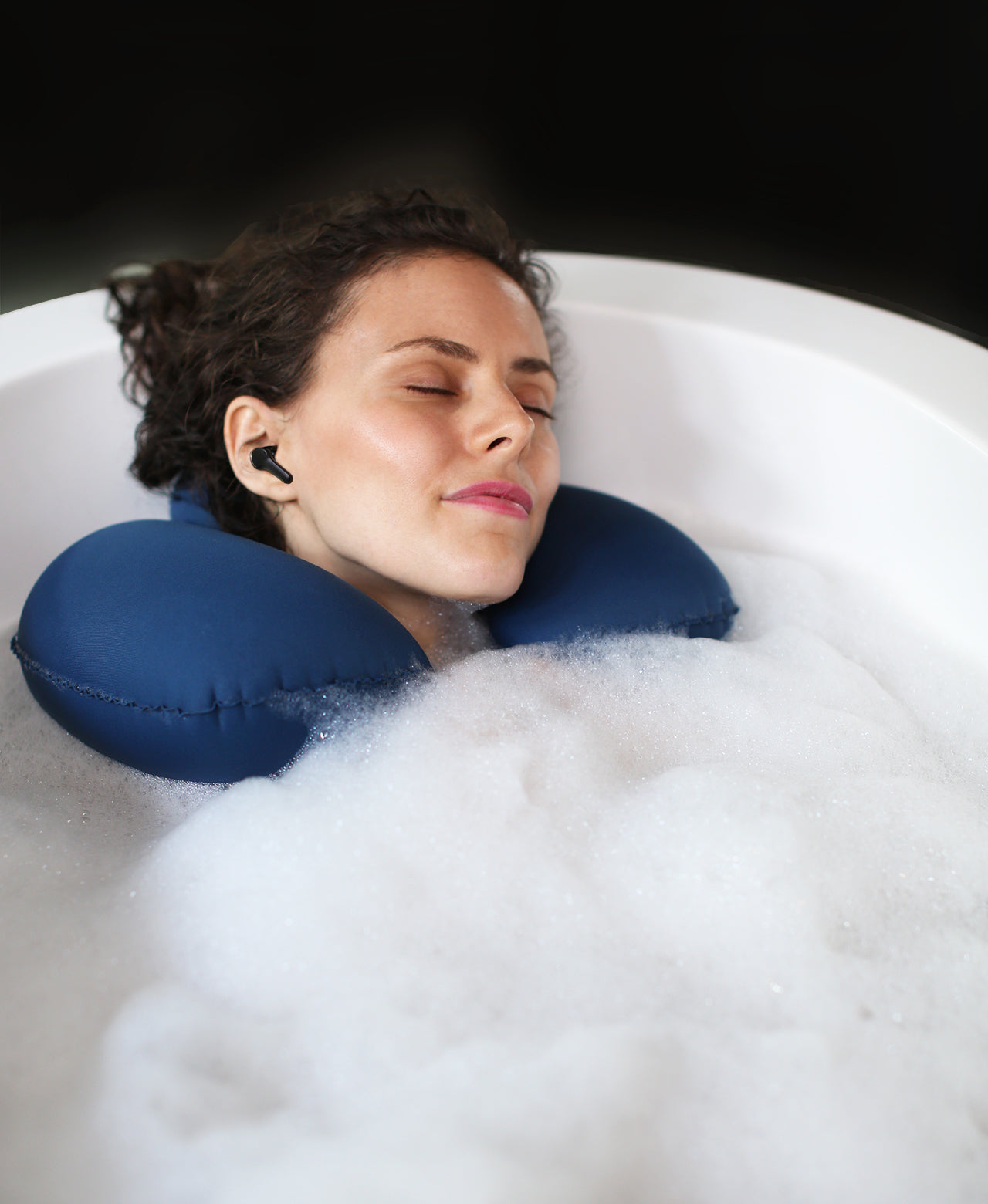 Person wearing BathSonic earbuds while resting in a foaming bath soak with a blue Head Floater snapped around their neck.